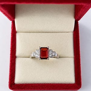 Puede incluir: Un anillo de tono plateado con una piedra central rectangular de color rojo intenso sujeta por cuatro garras. La banda está decorada a cada lado con tres pequeñas piedras transparentes de corte cuadrado en un patrón geométrico escalonado.