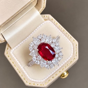 Puede incluir: Un anillo con una gran piedra central de rubí rojo de corte ovalado rodeada por un halo de múltiples diamantes blancos de corte pera y redondo. La banda está incrustada con pequeños diamantes redondos en un engaste de metal plateado.
