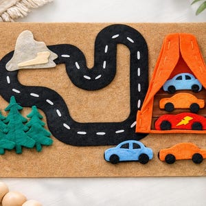 May include: A felt play mat featuring a black road with white dashed lines, green felt trees, and a mountain. An orange felt structure houses three colorful toy cars. Two additional toy cars are on the mat.