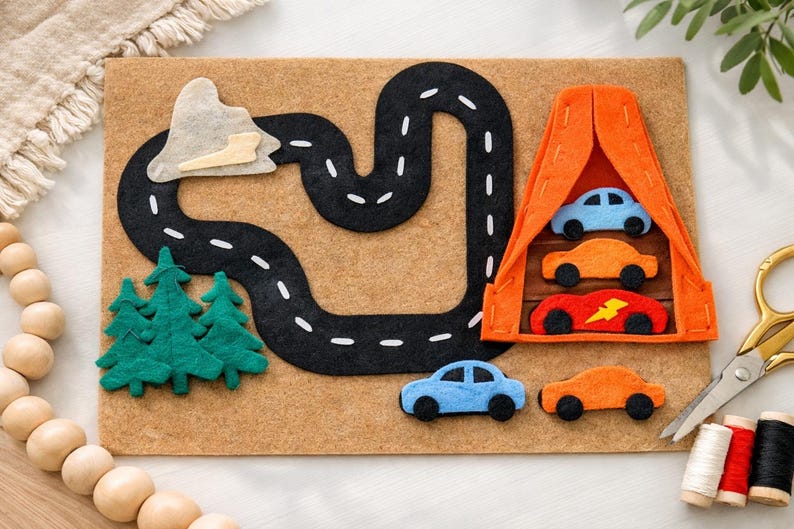 May include: A felt play mat featuring a black road with white dashed lines, green felt trees, and a mountain. An orange felt structure houses three colorful toy cars. Two additional toy cars are on the mat.