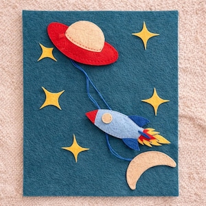 Puede incluir: Manualidad de fieltro con temática espacial que presenta un OVNI rojo y beige conectado por un hilo azul a un cohete azul con llamas rojas y amarillas. Estrellas amarillas y una luna creciente beige también están presentes.