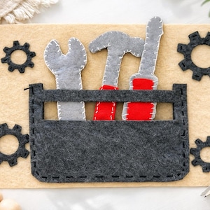 May include: A felt toolbox with gray tools and red accents sits on a tan background. The toolbox contains a wrench, hammer, and screwdriver. Black gear shapes are on the tan background. The tools are gray with white stitching.
