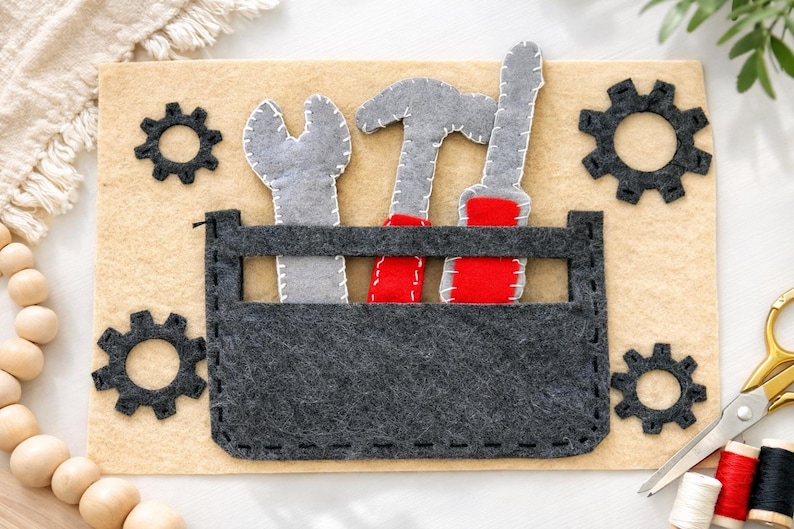 May include: A felt toolbox with gray tools and red accents sits on a tan background. The toolbox contains a wrench, hammer, and screwdriver. Black gear shapes are on the tan background. The tools are gray with white stitching.