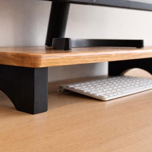 Peut inclure: Un support d'écran en bois avec un dessus brun clair et des pieds noirs. Le support est conçu pour surélever un écran d'ordinateur, avec un clavier placé en dessous. Le support est en bois et a un design simple et moderne.