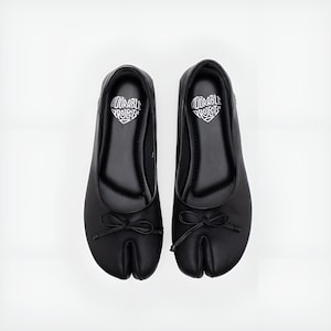 May include: A pair of black split-toe ballet flats. Each shoe has a small bow at the toe and the text "Adorable Projects" inside a heart-shaped design on the insole. The shoes are on a white background.