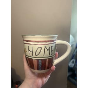 May include: Cream-colored ceramic mug with the word "HOME" in black lettering. The mug features a star and stripes design in red, brown, and blue. The handle is white.