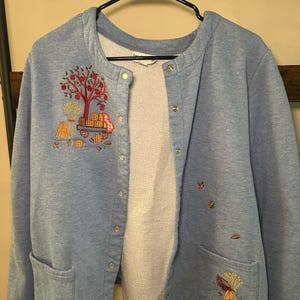 May include: Light blue button-up jacket with embroidered autumn-themed designs. Features a tree with red apples, a hay bale, and a wagon on the front. Additional embroidered leaves and a small house are on the right side. Includes two front pockets.