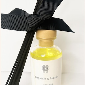 May include: A clear glass diffuser bottle with a gold cap, filled with yellow liquid. The label reads "Bergamot & Pepper" and "Diffuser." Black reeds and a black ribbon are tied around the neck. The bottle contains 100ml of liquid.