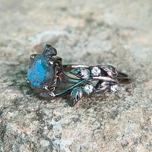 Könnte beinhalten: Ein silberner Ring mit einem großen, ovalen, blauen Edelstein. Der Ring hat ein Blatt- und Ranken-Design mit kleinen, klaren Edelsteinen. Der Ring ist vor einem strukturierten, grauen Hintergrund platziert.