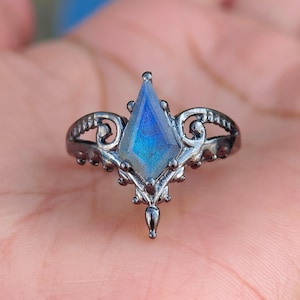 May include: A dark metal ring featuring a kite-shaped labradorite gemstone with blue iridescence. The band displays intricate scrollwork and filigree detailing, with a pointed setting that extends downward to a small metal bead at the base.