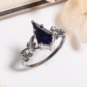 May include: A silver-toned ring featuring a kite-shaped dark blue sandstone center stone with sparkling inclusions. The band is designed with intricate metal leaf motifs and small, clear round-cut stones set along the sides of the setting.