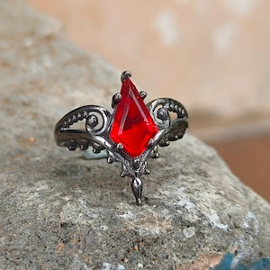 Puede incluir: Un anillo de tono gris oscuro con una piedra preciosa roja en forma de cometa en el centro. La banda de metal presenta intrincados adornos de volutas y detalles de cuentas, con la piedra roja sujeta por cuatro garras en un diseño simétrico.