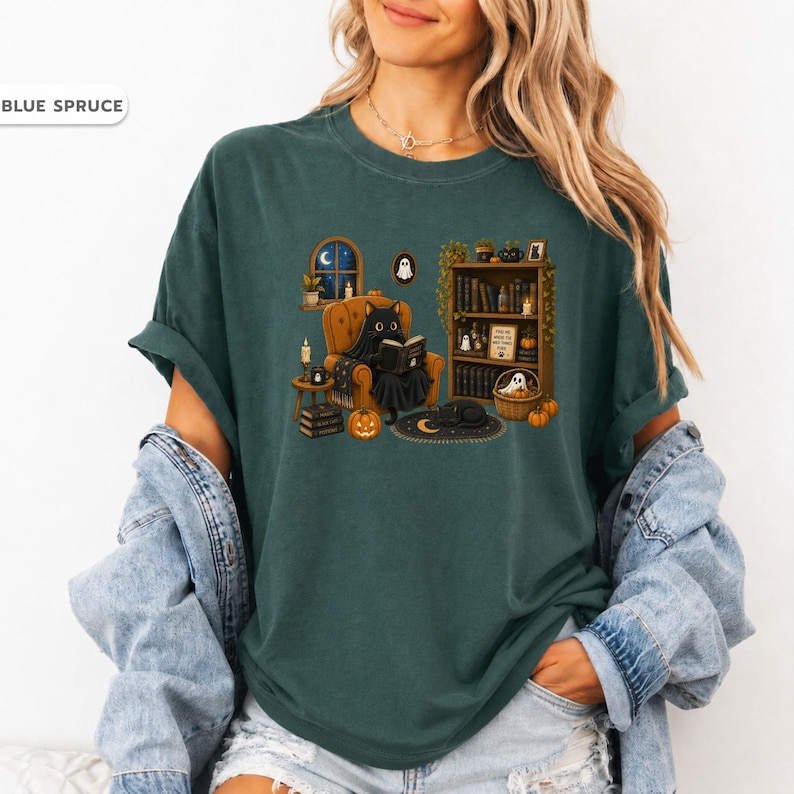 May include: A blue spruce t-shirt featuring a graphic of a black cat sitting in an orange armchair reading a book. The scene includes a bookshelf, a window with a night sky, pumpkins, and small ghost figures. The text "BLUE SPRUCE" is visible.