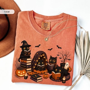 Op de afbeelding: Een roestoranje t-shirt met een grafische print van vier zwarte katten die boeken lezen, een pompoen, een spook en vleermuizen. Het shirt heeft een ronde hals en een Comfort Colors label in de nek. Een gouden ketting ligt bovenop het t-shirt.