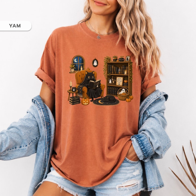 May include: A rust-colored t-shirt featuring a graphic illustration of a black cat sitting in an armchair reading a book. The scene includes a bookshelf, pumpkins, candles, and a small ghost. The text YAM is visible in the upper left corner.