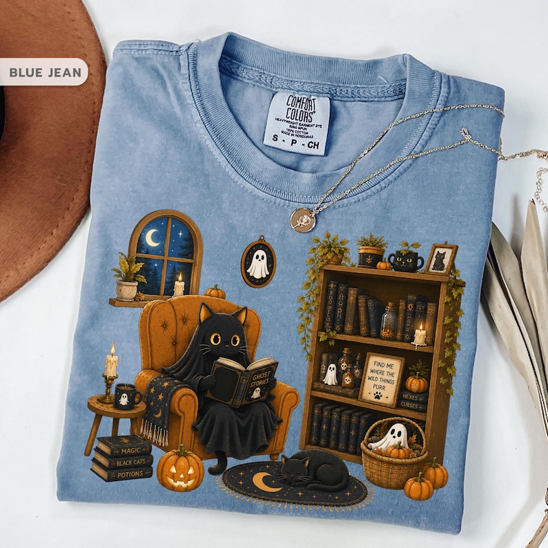 May include: A blue jean colored t-shirt featuring a graphic of a black cat sitting in an armchair reading a book titled Ghost Stories. The design includes a bookshelf, pumpkins, a ghost, a window with a crescent moon, and the text Find Me Where The Wild Things Purr.
