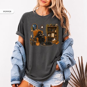 May include: A charcoal gray t-shirt featuring a graphic print of a black cat wearing a ghost costume while reading a book in an armchair. The scene includes a bookshelf, pumpkins, candles, and a crescent moon rug. The word PEPPER is printed in the corner.