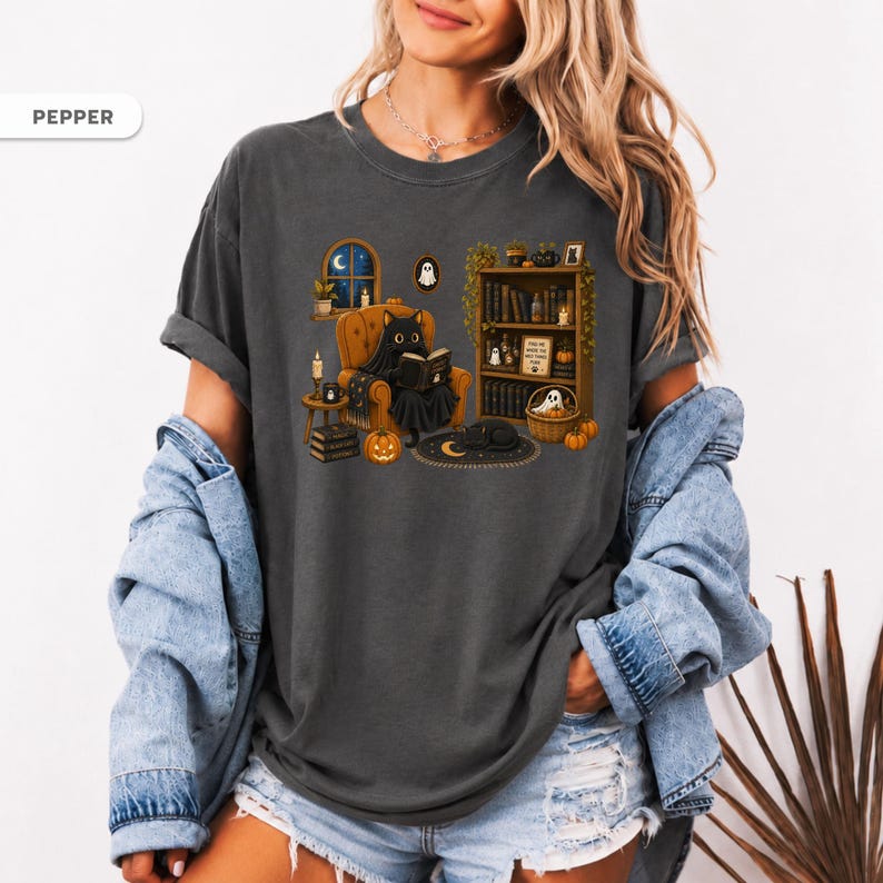 May include: A charcoal gray t-shirt featuring a graphic print of a black cat wearing a ghost costume while reading a book in an armchair. The scene includes a bookshelf, pumpkins, candles, and a crescent moon rug. The word PEPPER is printed in the corner.