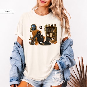 May include: An ivory t-shirt featuring a graphic of a black cat sitting in an orange armchair reading a book. The scene includes a bookshelf, a window with a night sky, pumpkins, a small ghost, and a black cat sleeping on a crescent moon rug.