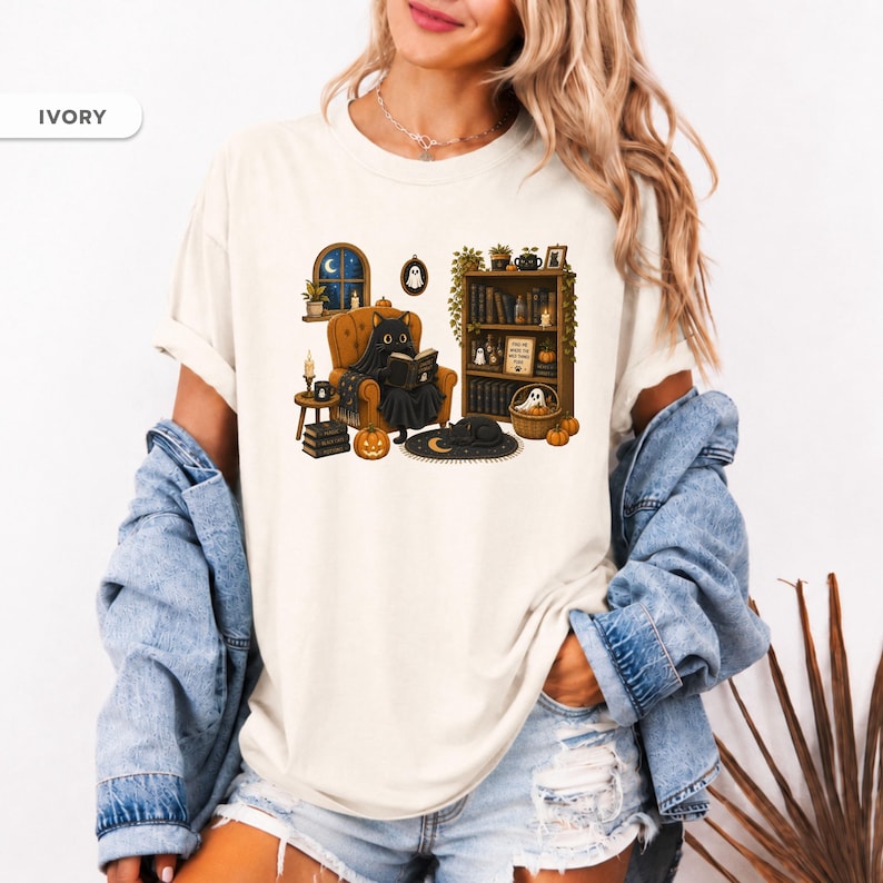 May include: An ivory t-shirt featuring a graphic of a black cat sitting in an orange armchair reading a book. The scene includes a bookshelf, a window with a night sky, pumpkins, a small ghost, and a black cat sleeping on a crescent moon rug.