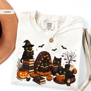 Op de afbeelding: Een ivoorkleurig t-shirt met een afbeelding van vier zwarte katten die boeken lezen in een Halloween-setting. Het ontwerp bevat pompoenen, vleermuizen, een kleine boom en boeken met de titels Spells, Potions, Black Magic, Curses en Pumpkin Tales.