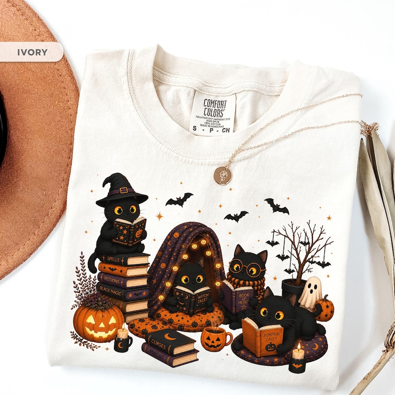 Op de afbeelding: Een ivoorkleurig t-shirt met een afbeelding van vier zwarte katten die boeken lezen in een Halloween-setting. Het ontwerp bevat pompoenen, vleermuizen, een kleine boom en boeken met de titels Spells, Potions, Black Magic, Curses en Pumpkin Tales.