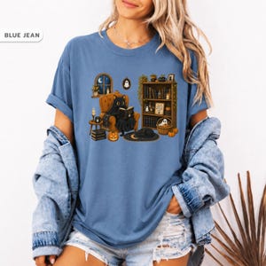 May include: A blue jean colored t-shirt featuring a graphic print of a black cat reading a book in an armchair, surrounded by a bookshelf, pumpkins, a ghost, and a window. The text on the bookshelf sign reads "YES WE HAVE NO NEW BOOKS".