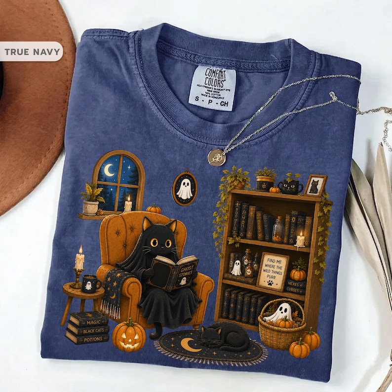 May include: A navy blue t-shirt featuring a graphic of a black cat sitting in an orange armchair reading a book titled Ghost Stories. The design includes a bookshelf, pumpkins, candles, a window with a crescent moon, and a small ghost illustration.