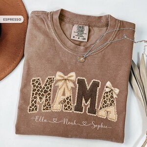 Peut inclure: Un t-shirt marron Comfort Colors avec le mot "MAMA" dans un design décoratif. Les lettres ont un imprimé léopard et une texture de toile de jute, avec des nœuds. Les noms "Ella", "Noah" et "Sophie" sont écrits en dessous.
