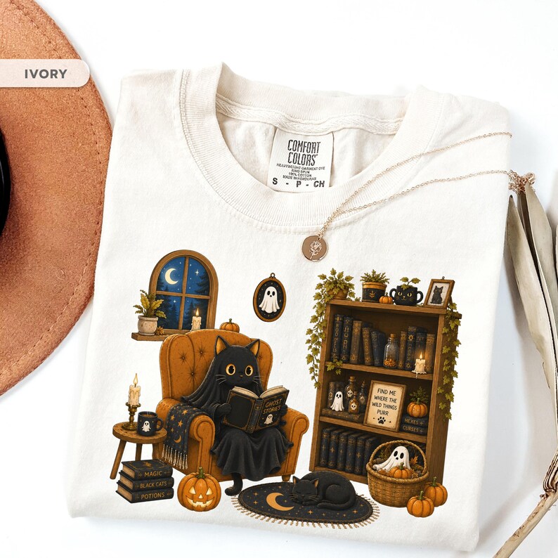 May include: An ivory t-shirt featuring a graphic of a black cat sitting in an orange armchair reading a book. The design includes a bookshelf, pumpkins, a ghost, candles, and a window with a night sky. The shirt has a Comfort Colors brand label.