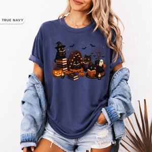 Op de afbeelding: Een marineblauw t-shirt met korte mouwen en een grafische print van zwarte katten die boeken lezen, pompoenen en vleermuizen. Het ontwerp bevat verschillende katten met heksenhoeden, stapels boeken, een klein spookje en brandende kaarsen als een gedetailleerde Halloween-illustratie op de voorzijde.