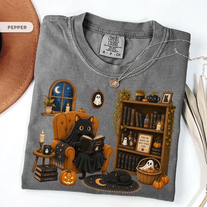 May include: A pepper-colored t-shirt featuring a graphic of a black cat wearing a ghost sheet and reading a book titled Ghost Stories in an orange armchair. The design includes a bookshelf, a window with a moon, pumpkins, and a ghost.