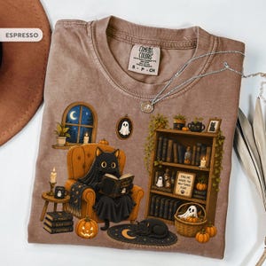 May include: An espresso-colored t-shirt featuring a graphic of a black cat sitting in an orange armchair reading a book titled Ghost Stories. The design includes a bookshelf, pumpkins, a ghost, a window with a moon, and the text Find Me Where The Wild Things Purr.