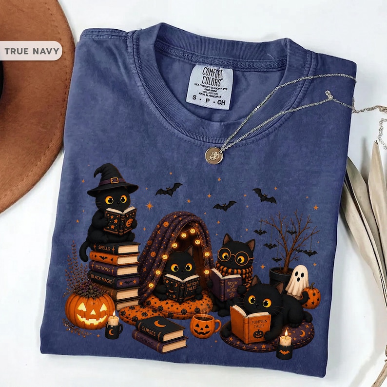 Op de afbeelding: Een marineblauw t-shirt met een afbeelding van vier zwarte katten die boeken lezen, omringd door pompoenen, vleermuizen en een klein spookje. Het t-shirt heeft een ronde hals, een Comfort Colors-label en een zilveren ketting.