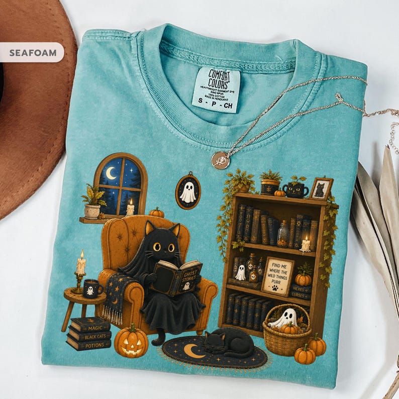 May include: A seafoam green t-shirt featuring a graphic of a black cat wearing a ghost sheet while reading a book in an armchair. The design includes a bookshelf, pumpkins, candles, and a cat sleeping on a rug. The text reads "Find me where the wild things purr".