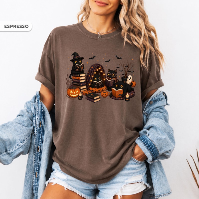 Op de afbeelding: Een espresso-kleurig t-shirt met een grafische print van zwarte katten die boeken lezen, pompoenen, een spook en vleermuizen. De tekst ESPRESSO staat in de linkerbovenhoek. Het ontwerp bevat oranje, zwarte en bruine tinten op een donkerbruin shirt.