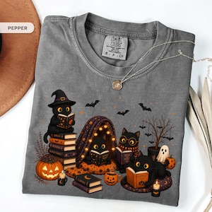 Op de afbeelding: Een peperkleurig t-shirt met een afbeelding van vier zwarte katten die boeken lezen. Het ontwerp bevat een stapel boeken, een pompoen, een klein spookje, een boom met hangende vleermuizen en teksten op boekomslagen zoals Spells, Black Magic en Pumpkin Tales.