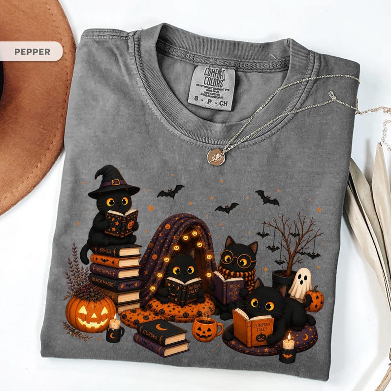 Op de afbeelding: Een peperkleurig t-shirt met een afbeelding van vier zwarte katten die boeken lezen. Het ontwerp bevat een stapel boeken, een pompoen, een klein spookje, een boom met hangende vleermuizen en teksten op boekomslagen zoals Spells, Black Magic en Pumpkin Tales.