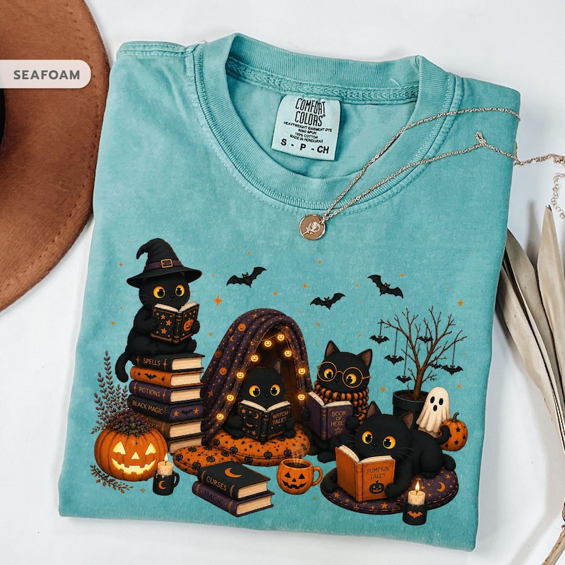 Op de afbeelding: Een zeegroen t-shirt met een grafische print van vier zwarte katten die boeken lezen in een Halloween-setting. Het ontwerp bevat pompoenen, een spook, vleermuizen en een kleine boom. Het t-shirt heeft een Comfort Colors label in de nek.