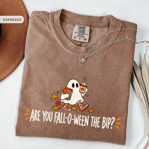 May include: An espresso-colored t-shirt featuring a graphic of a ghost holding books and a coffee cup, surrounded by autumn leaves. The text reads, "ARE YOU FALL-O-WEEN THE BIP?" The shirt has a Comfort Colors label at the neck and a crew neckline.
