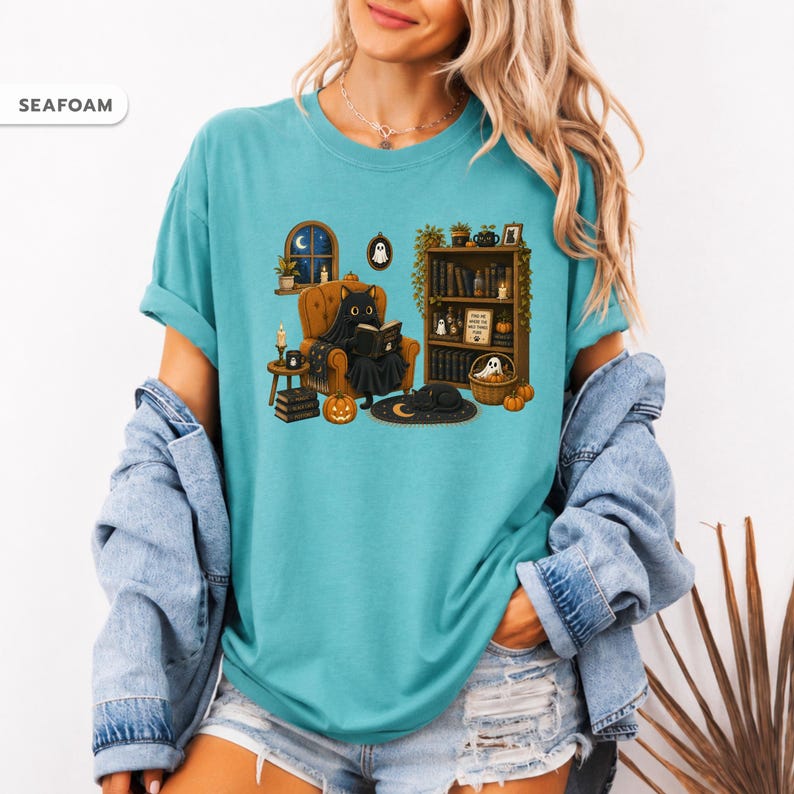 May include: A seafoam green t-shirt featuring a graphic illustration of a black cat sitting in an armchair reading a book. The scene includes a bookshelf, pumpkins, candles, and a ghost. The text "SEAFOAM" appears in a small white box in the corner.