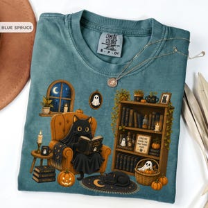 May include: A blue spruce t-shirt featuring a graphic of a black cat wearing a ghost sheet while reading a book in an orange armchair. The scene includes a bookshelf, pumpkins, candles, and a framed sign that reads Find me where the wild things purr.