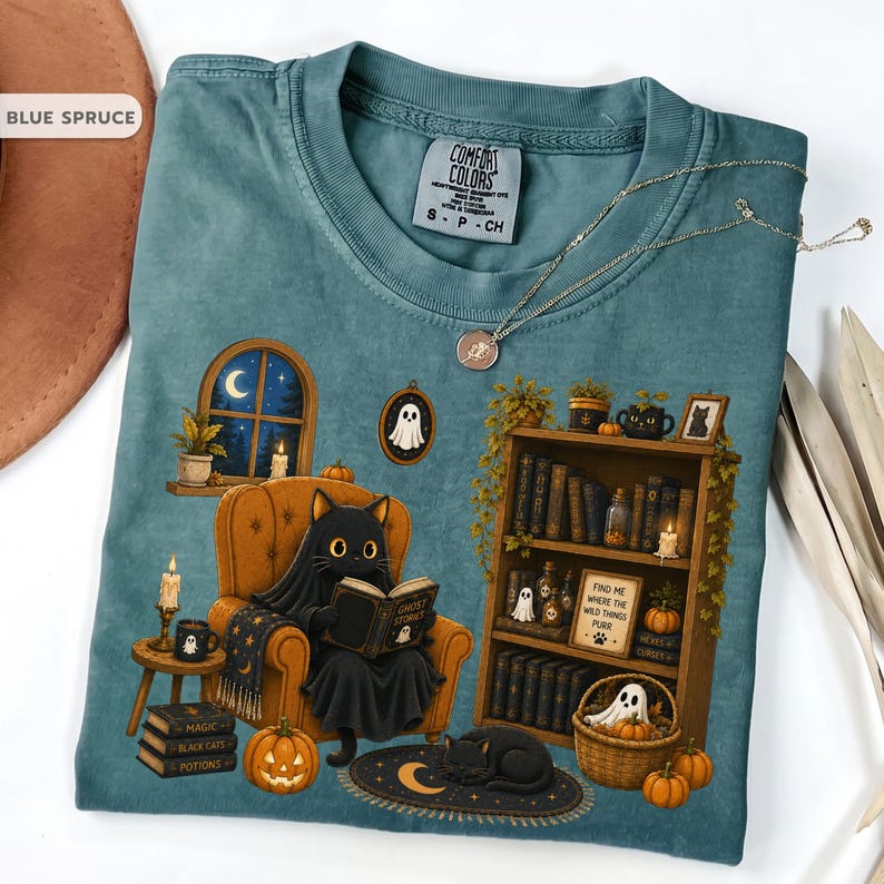 May include: A blue spruce t-shirt featuring a graphic of a black cat wearing a ghost sheet while reading a book in an orange armchair. The scene includes a bookshelf, pumpkins, candles, and a framed sign that reads Find me where the wild things purr.