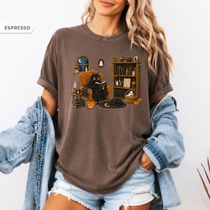 May include: An espresso-colored t-shirt featuring a graphic of a black cat sitting in an orange armchair reading a book. The scene includes a bookshelf, pumpkins, a crescent moon rug, and a window with a night sky. The text ESPRESSO appears in the corner.