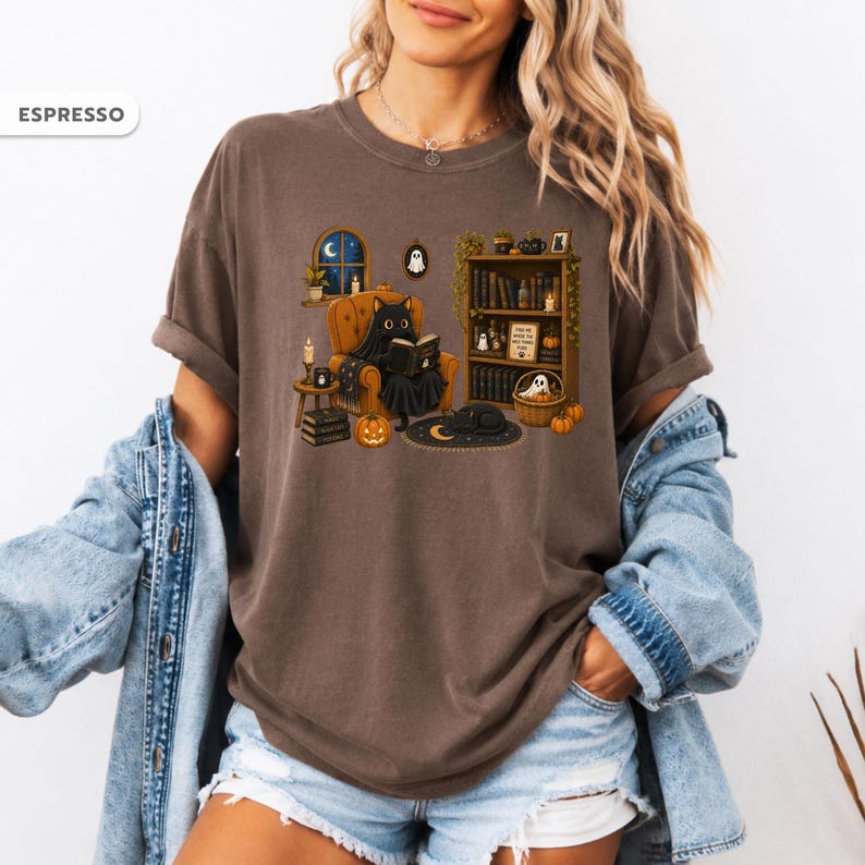 May include: An espresso-colored t-shirt featuring a graphic of a black cat sitting in an orange armchair reading a book. The scene includes a bookshelf, pumpkins, a crescent moon rug, and a window with a night sky. The text ESPRESSO appears in the corner.