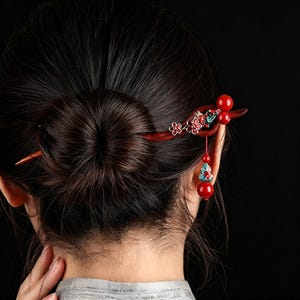 Könnte beinhalten: Eine dunkelrote Haarnadel aus Holz mit einem dekorativen Blumenmotiv mit roten und türkisfarbenen Emaille-Akzenten, zwei glatten roten Kugeln und einem hängenden Anhänger, der aus einer kleinen türkisfarbenen und roten Perle und einer roten Kugel besteht.