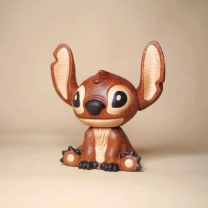 K&ouml;nnte beinhalten: Eine Holzfigur von Stitch, der Disney-Figur, in sitzender Position. Die Figur ist haupts&auml;chlich braun mit helleren Holzdetails an Ohren und Brust. Sie hat gro&szlig;e schwarze Augen und eine schwarze Nase, wobei die Ohren nach oben zeigen.