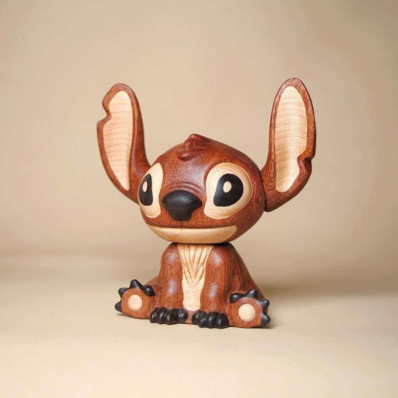 K&ouml;nnte beinhalten: Eine Holzfigur von Stitch, der Disney-Figur, in sitzender Position. Die Figur ist haupts&auml;chlich braun mit helleren Holzdetails an Ohren und Brust. Sie hat gro&szlig;e schwarze Augen und eine schwarze Nase, wobei die Ohren nach oben zeigen.