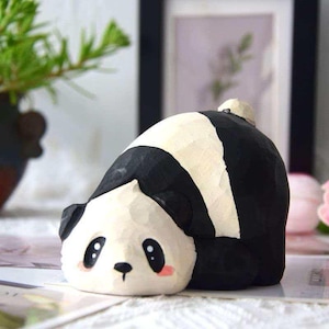 May include: A hand-carved wooden panda figurine in a lying down pose. The panda has a black and white color scheme with pink cheeks and large, expressive eyes. The figurine is on a table with a blurred background.