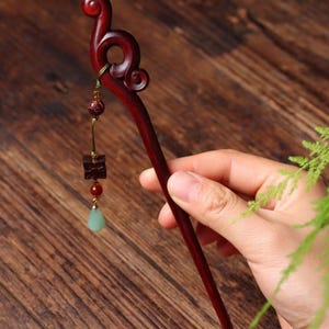 May include: A dark reddish-brown wooden hair stick featuring a carved scroll-shaped top. A gold-toned metal chain hangs from the top, adorned with a small round bead, a square wooden charm, a tiny red bead, and a pale green teardrop-shaped pendant.